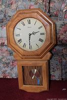 Full view of the wooden clock, showing octagonal wooden frame, the white clock face with Roman numerals, and the wooden lower case with glass exposing the pendulum and battery.
