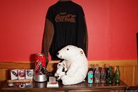 Overview of Coca Cola memorabilia including polar bear plush dolls, the jacket, large red can lamp, various collectibles and bottles on tabletop against red wall.