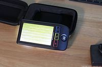 Handheld magnifier powered on displaying magnified wood grain next to open zippered carrying case