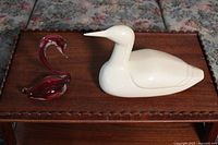 Two decorative bird figurines on a wooden surface: red glass swan and white ceramic loon, shown side by side.