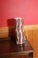 Front and side views of a tall cylindrical glass vase with abstract brown brushstroke designs on a white base. The vase is placed on a wood surface with a red wall background.