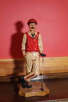 Full front view of the golf figure statue showing detailed red vest, beige pants with stripes, and golf club on wooden base.
