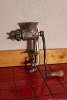 Side view of vintage metal manual meat grinder, showing brand and model embossing, hand crank with wooden handle, and clamp attachment.