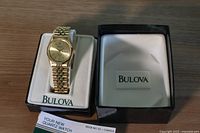 Bulova gold-tone men's wristwatch displayed in original white and black box with Bulova logo and owner's guide booklet included.