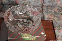 Clear glass bowl with detailed embossed peacock feather and leafy floral patterns around the rim, resting on the original Flamenca box showing a large colorful peacock feather illustration and brand name.