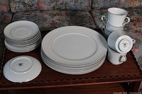 White porcelain set of plates, tea cups, and saucers stacked on wooden table with floral couch in background. Includes 5 large plates on the bottom, 2 dessert plates on top, and 6 saucers next to stacked tea cups.