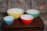 Set of four Pyrex mixing bowls displayed on a wooden surface with floral background. Bowls in blue, red, green, and yellow, arranged from smallest to largest.