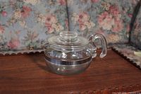 Side view of clear glass Pyrex tea pot showing silver band and curved handle.