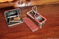 Two vintage safety razor sets in poor condition displayed on wood surface. Includes Gillette razor in black hinged case and Rolls razor with chrome parts and metal case with removable lid.