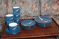 Six blue tea cups stacked, six saucers stacked, six dessert plates stacked, each featuring white shrimp design on blue background with gold rims, displayed on wooden table with floral sofa background.