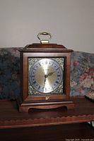 Front view of wooden mantel clock showing clock face with Roman numerals and decorative metal corners.