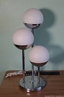 Photo showing front view of the table lamp with three white spherical shades on chrome stems and round chrome base.