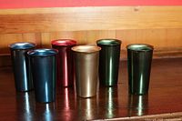 Six vintage anodized aluminium drinking glasses in assorted metallic colors arranged standing on a wooden table.