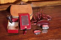 Vintage maroon Norelco Sportsman battery razor with power cord, red mirror, spare parts in metal box, and brown zippered carrying case on wooden surface.