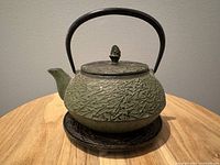 Full view of green cast iron teapot with black handle and lid, sitting on trivet.