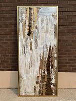 Full view of large framed abstract painting leaning against a brick wall, showing tall vertical textured brush strokes in muted and metallic tones.