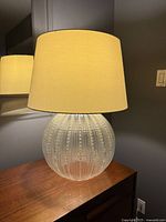 Front-view photo of the large glass table lamp showing its textured spherical clear glass base and beige lampshade, lit up to display warm light and fabric texture.