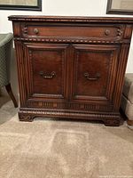 Front view of carved wooden cabinet with drawer above double doors adorned with metal handles and carved trim.