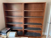 Two medium brown wooden book shelves side by side, each with five shelves lined with black shelf liners. Visible minor wear and some dust on shelves.