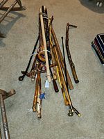 Photo showing approximately 14 wooden canes of various sizes and shapes, some with curved handles, others straight with rounded or detailed ends, arranged on carpet floor.
