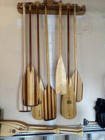 Ten wooden canoe paddles displayed—seven solid wood and three laminated striped—hanging on a wall rack with two additional paddles on the floor.