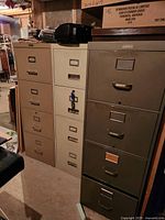 Photo showing three filing cabinets in a basement or storage area. Beige, off-white, and green filing cabinets arranged side by side.