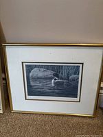 Close-up of one framed loon print showing loon on water, gold frame, white mat, signed by artist Don Li-Leger