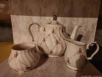 Photo showing a white porcelain teapot, sugar bowl, and creamer. Each has pink rose decoration and gold diamond lattice detailing.