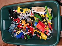 Top view of green bin containing assorted die-cast toy vehicles and a wooden train track piece