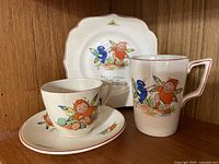 Photo showing three ceramic nursery items: a small cup with saucer and a mug, plus a decorative plate, all decorated with colorful gnome and insect illustrations and dated circa 1930.