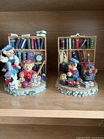 Two ceramic teddy bear figurines in a library setting with bookshelves and toys.