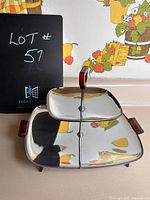 Three-section stainless steel serving tray with brown resin handles, footed base, and central carrying handle, photographed against fruit-themed background.