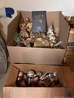 Two cardboard boxes filled with Christmas decorations and ornaments. The top box features gold-coloured angels and a gold Christmas tree with other gold-toned decorations. The bottom box contains various coloured Christmas baubles.