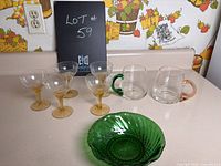 Photo showing four small wine glasses with yellow stems, two glass mugs with coloured handles, and a green depression glass bowl on a counter.