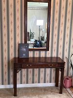 Hall table and mirror displayed against wallpapered wall. The table is wooden with brass handles and has a polished finish. The mirror is rectangular with a carved wooden frame matching the table.