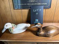 Photo of two wooden duck sculptures on a cabinet, one white painted with floral decorations, the other natural wood finish.