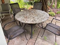 Photo of round stone look patio table with visible side damage and four mesh metal frame patio chairs arranged around it on a concrete patio.