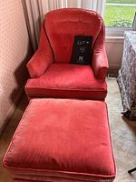 Front view of the pink upholstered chair and ottoman on casters in a room with carpet and near a window