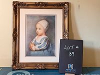 Full frontal photo of the framed print with an ornate gold frame and a child dressed in traditional blue and white clothing holding a rounded object.