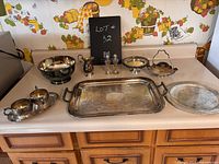 Photo of assortment of silver plated items showing large rectangular tray, various bowls, a pitcher, salt and pepper shakers, and other small serving pieces on a kitchen counter with vintage wallpaper background.