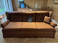 Photo showing front and left side of the brown fabric three-seater sofa with tufted backrest and three seat cushions, along with two bolster pillows.