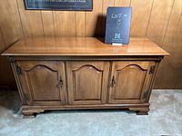 Front view of a traditional wooden cabinet with three paneled doors, brown wood finish, metal hinges and hardware, standing on decorative feet.