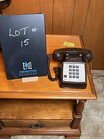 Brown touch-tone telephone sitting on a wooden side table next to a black sign with lot number.