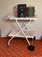 Full view of vintage bar cart displaying green leather ice bucket and metal cocktail shaker on top shelf, showing crossbar metal frame, two shelves with marble-like surface, two large black wheels and two caster wheels.