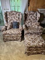 Two brown and beige floral wingback chairs and one matching ottoman shown from front, highlighting upholstery, shape and condition.