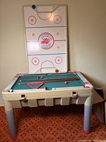 Full view of games table with air hockey surface upright, displaying air hockey playing area, pool cues, and balls on the green pool table surface below.