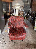Front view of vintage salon style hair dryer chair featuring red and black floral patterned upholstery, padded armrests, and dome hair dryer hood with attached cord.
