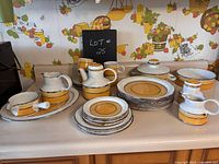 Full view of entire Midwinter dish set on kitchen counter showing arrangement of plates, cups, and serving pieces under 'LOT 25' sign