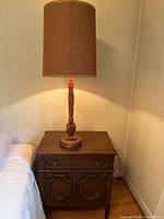 Vintage wooden table lamp on top of a wooden night stand next to a bed, lamp is lit, showing turned wood stem and fabric shade