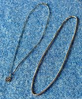 Photo showing both 925 sterling silver choker chains laid side by side on a blue fabric background, one chain has a circular H-shaped pendant, the other is a thicker box link style without pendant.